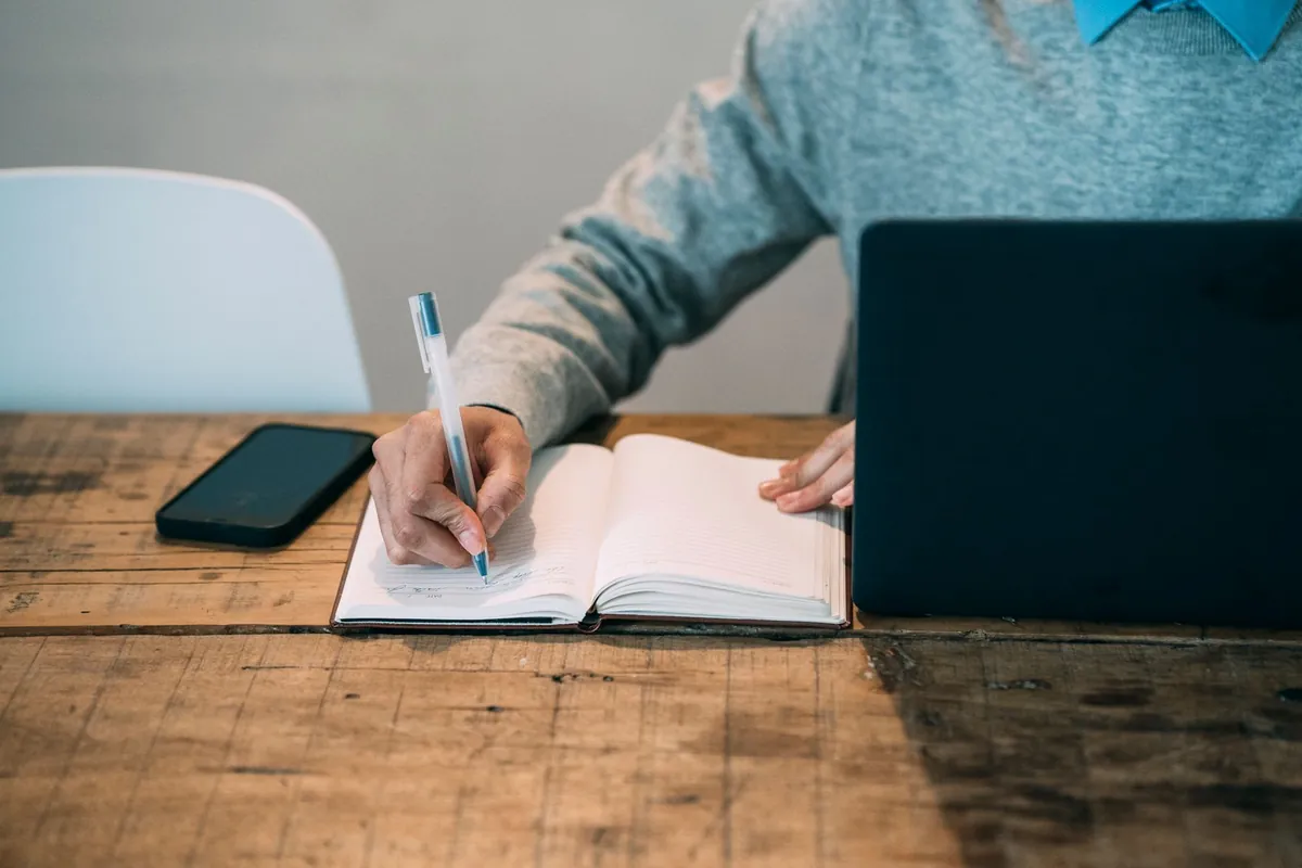 Person writing notes in a notebook beside a laptop while drafting a cover letter